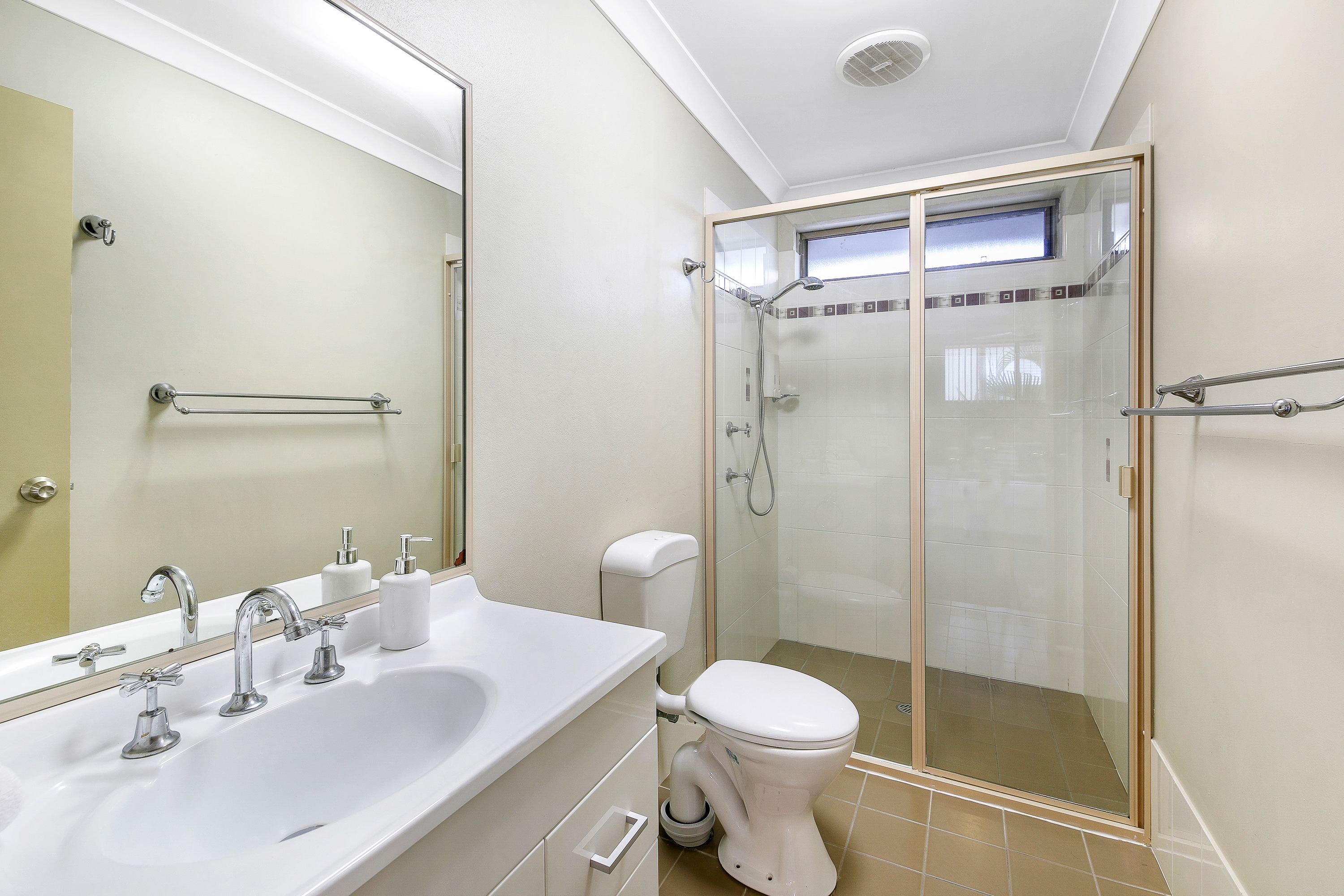 Un baño moderno con lavamanos, espejo y retrete blancos. Incluye una zona de ducha con mampara de cristal y una ventana para luz natural.
