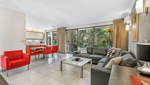 Un salón moderno con un sofá, sillas rojas, una mesa de centro de vidrio y vistas al exterior a través de amplios ventanales.