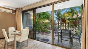 Una habitación de hotel moderna con un separador de vidrio, una mesa de comedor con sillas y vistas a un área exterior ajardinada con palmeras y una piscina.