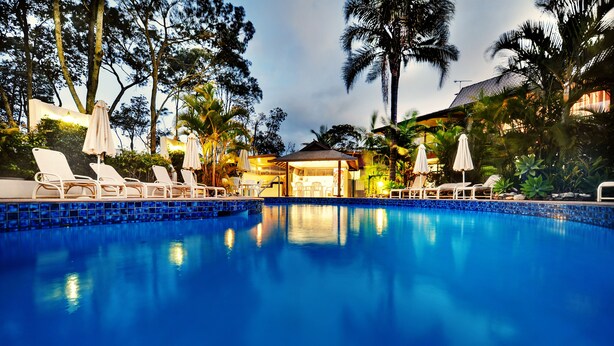 Un área de piscina con sillones de descanso y sombrillas, rodeada de palmeras y un edificio con tejado de baldosas.