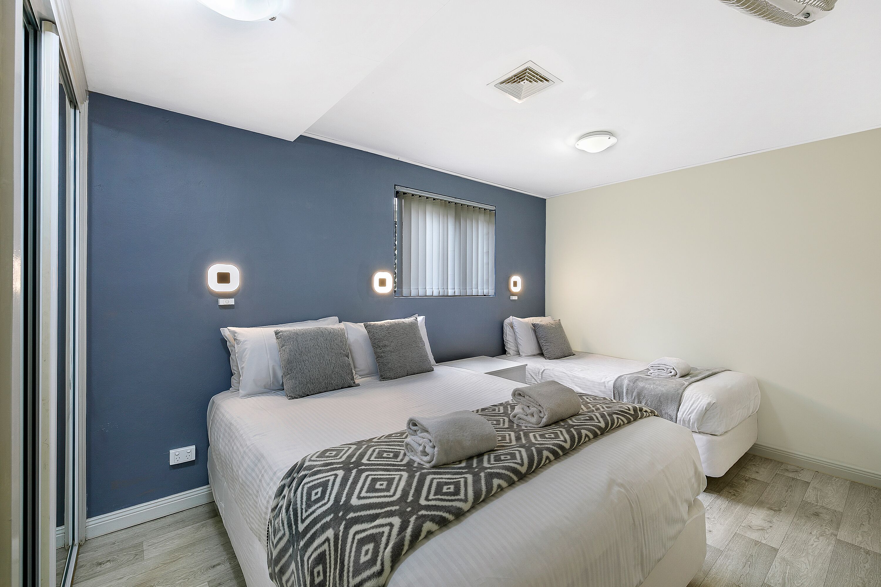 A bedroom with two beds, a patterned throw blanket, and wall-mounted lights.