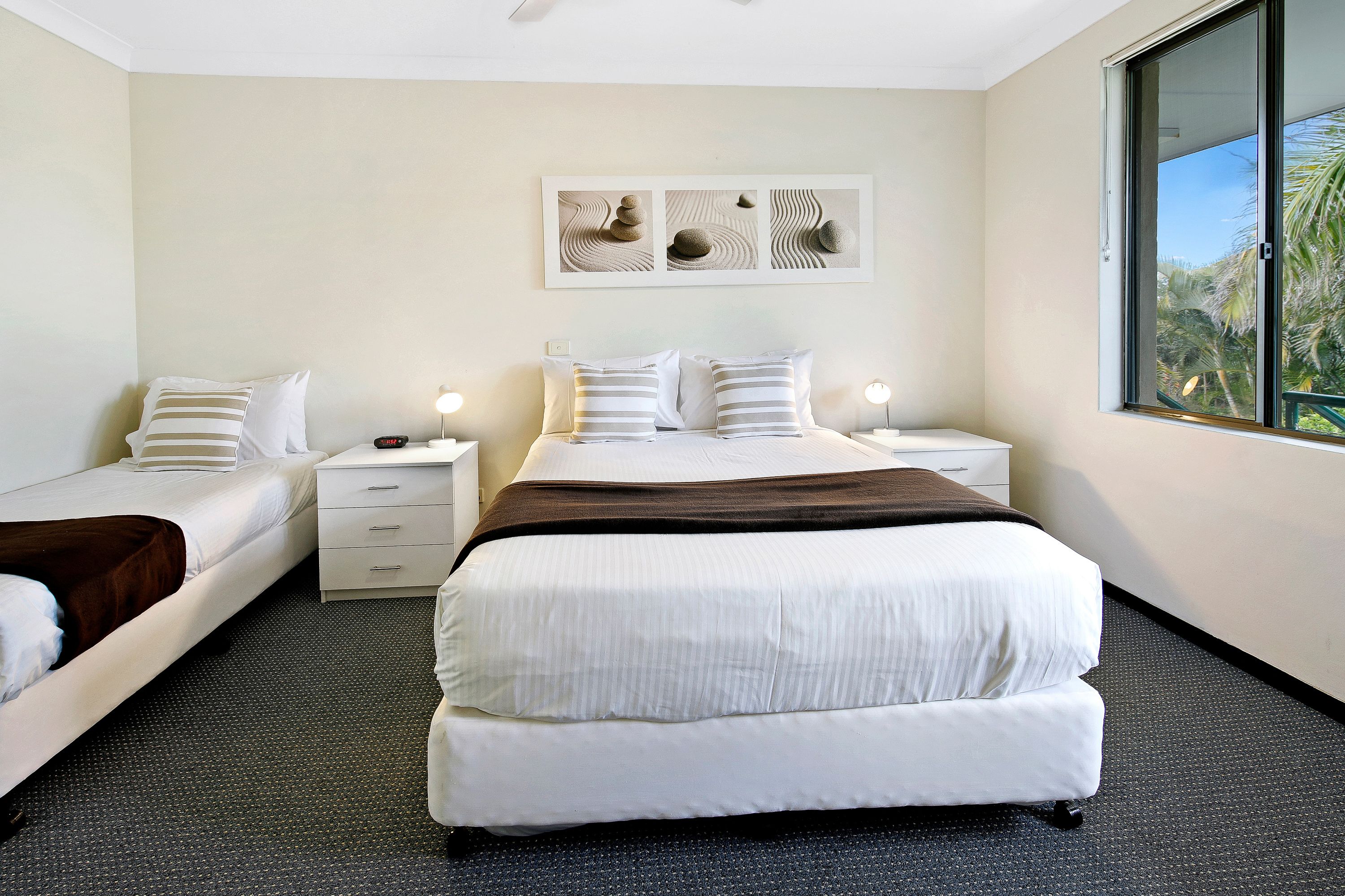 Habitación de hotel con una cama grande, dos camas más pequeñas, una mesita de noche con una lámpara, una obra de arte en la pared y una ventana con vistas a palmeras.