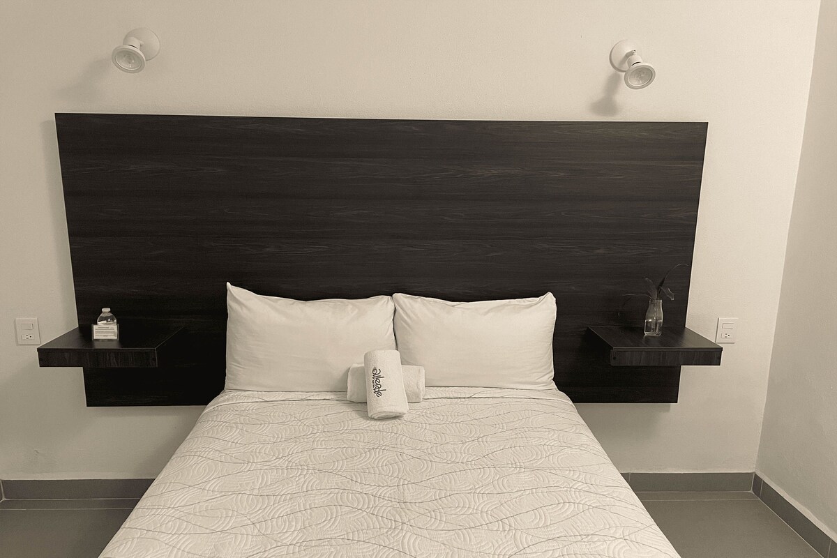 A hotel room with a dark wood headboard, two white pillows, a rolled-up towel, and two bedside tables with small items.