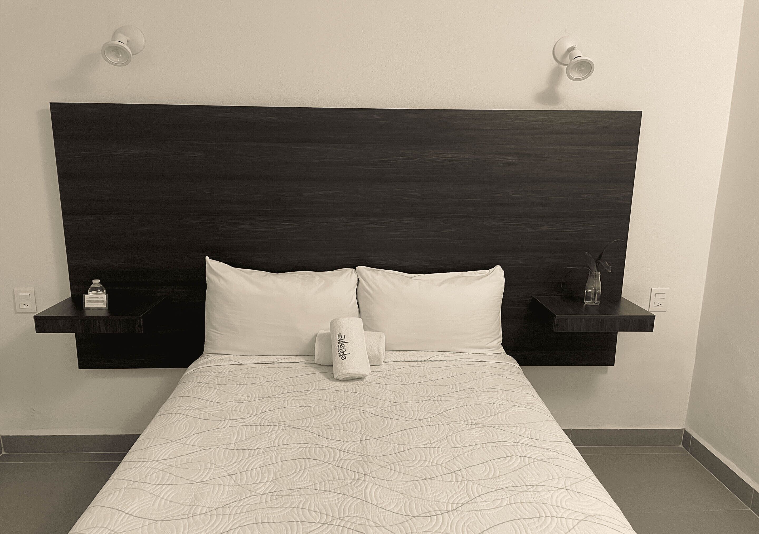 A hotel room with a dark wood headboard, two white pillows, a rolled-up towel, and two bedside tables with small items.