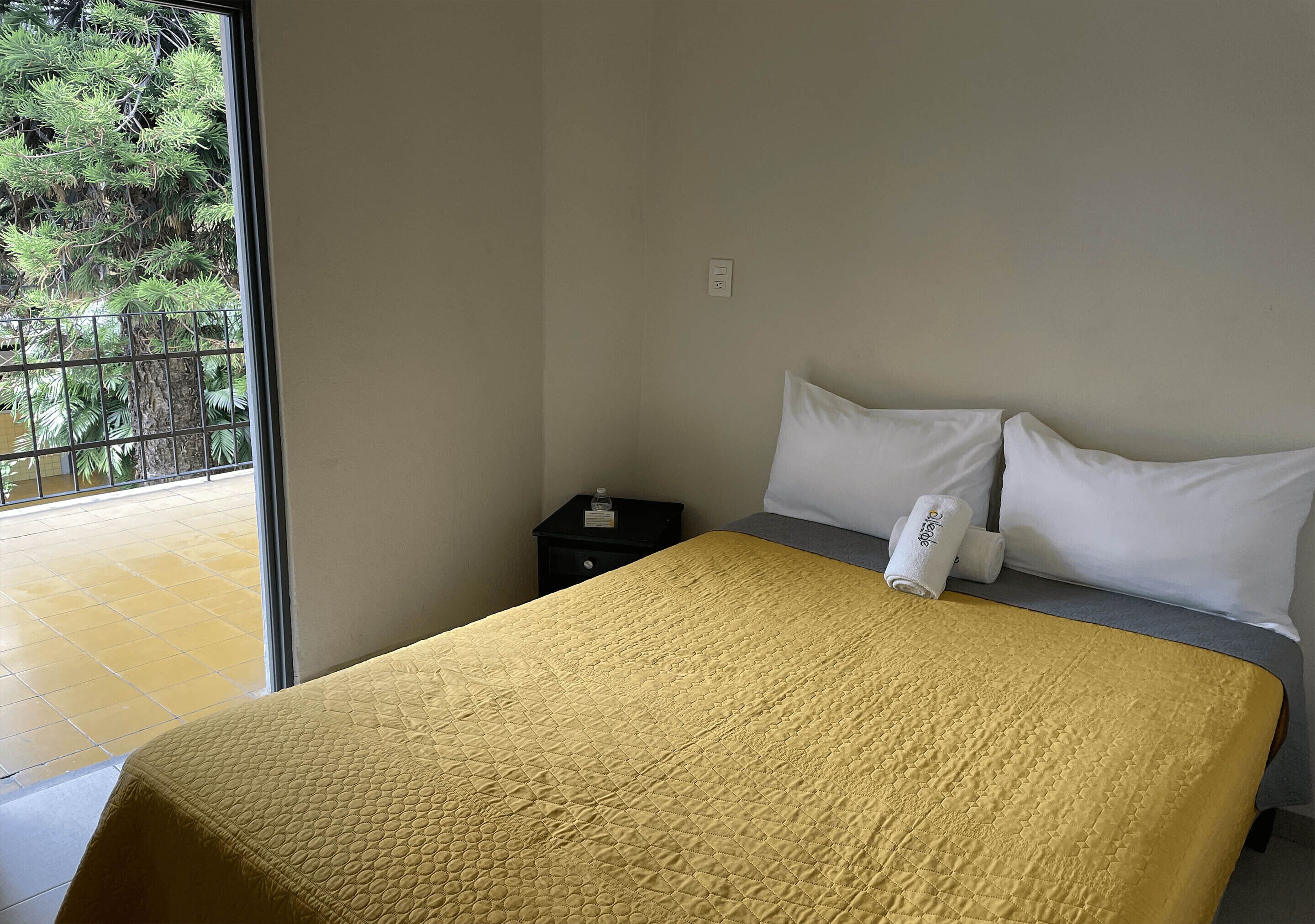 A hotel room with a bed, a nightstand, a window with a view of trees, and a tiled floor.
