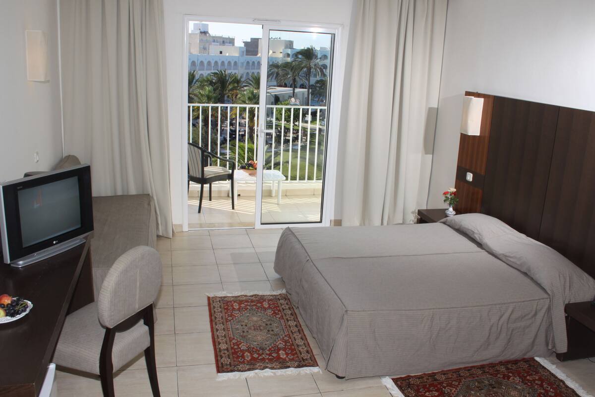 Habitación de hotel con cama, televisión, escritorio y silla. Balcón con vistas a palmeras y edificios.