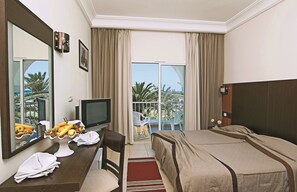 A hotel room with a bed, a desk with a television, a chair, a window with curtains, and a view of palm trees. - Primasol El Mehdi (Mahdia)