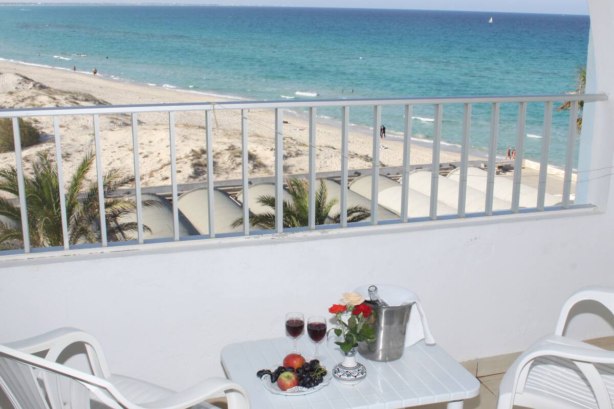 Un balcón con una mesa para dos, con vista a una playa donde se ven gente y un velero.