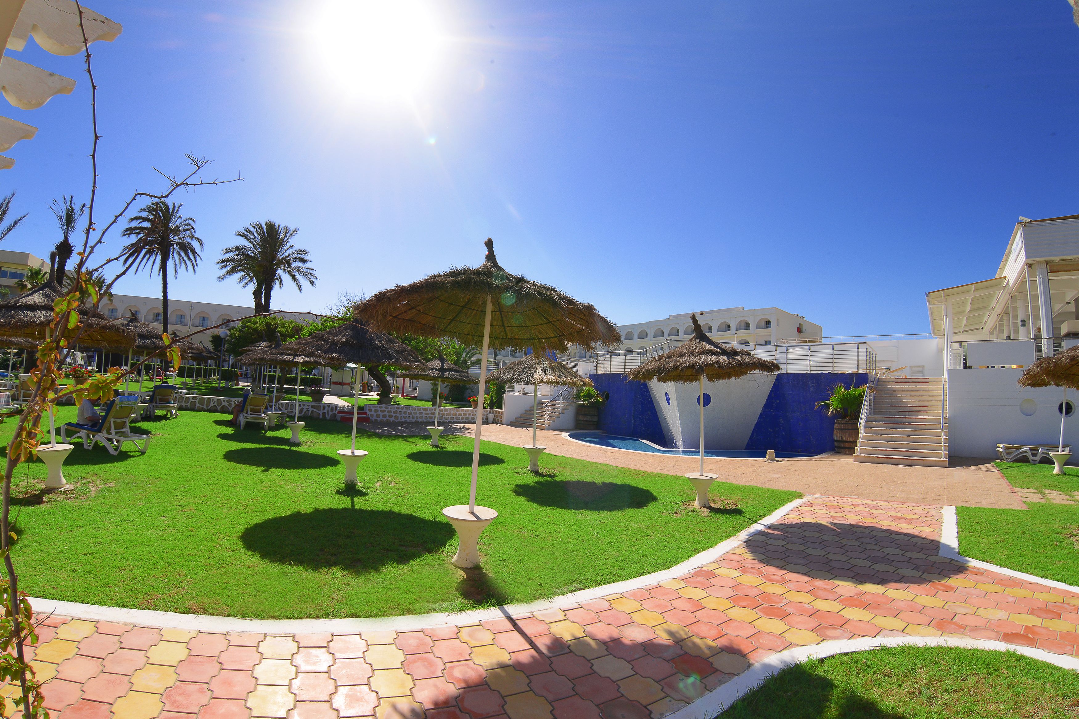 A courtyard with a pool, palm trees, and a thatched-roof structure.