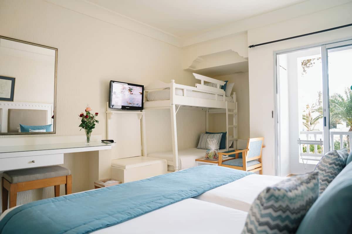 A room with a bunk bed, a desk with a mirror, a television, a chair, and a window.
