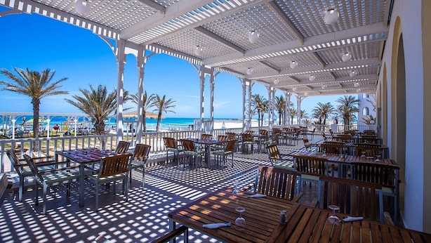 An outdoor dining area with tables and chairs, a white pergola structure, and a view of the beach.