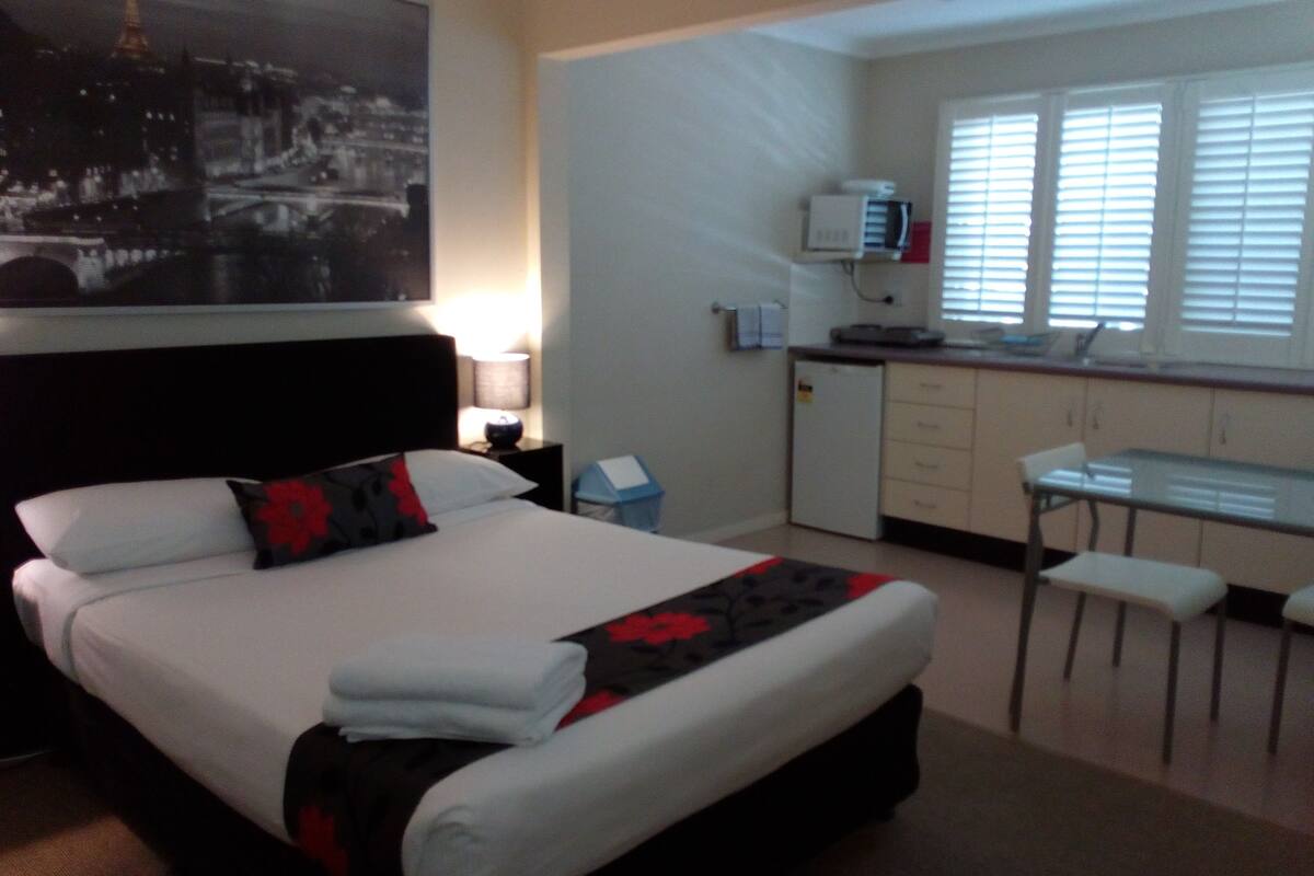 Habitación de hotel con cama, escritorio, silla, kitchenette pequeño y ventana con persianas.