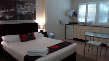 A hotel room with a bed, a desk, a chair, a small kitchenette, and a window with blinds.