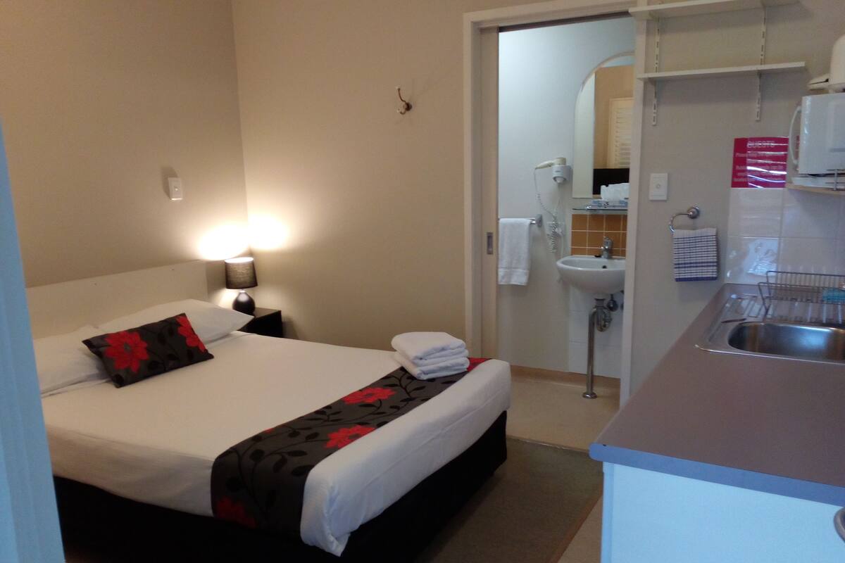 Habitación de hotel con cama, mesita de noche, lámpara y una pequeña kitchenette con lavabo y encimera.