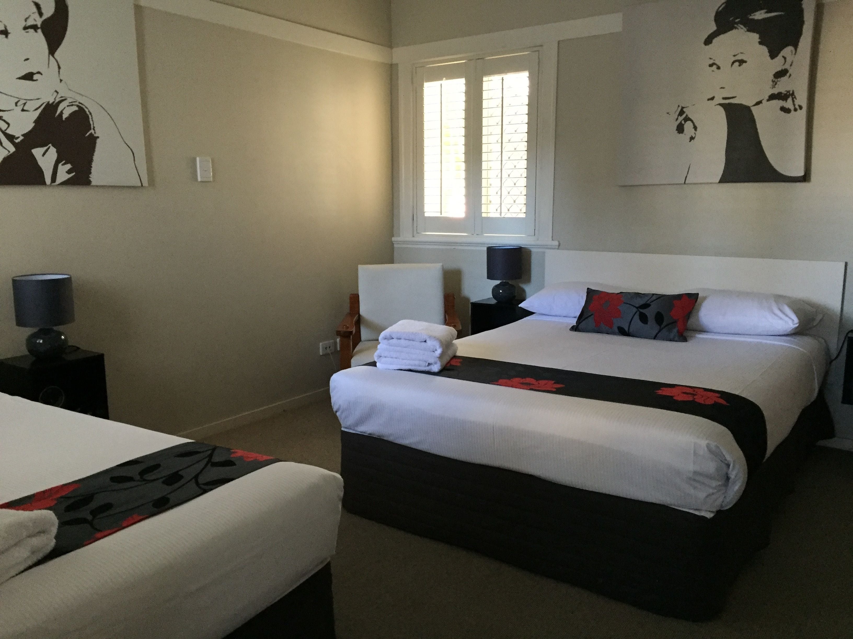 A hotel room with two beds, a window with blinds, a bedside table with a lamp, and artwork on the walls.