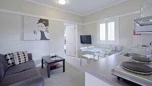 A compact living space with a sofa, a small table, a TV, a kitchen area with a stove, and a window with blinds. - Brisbane Street Studios (Annerley)