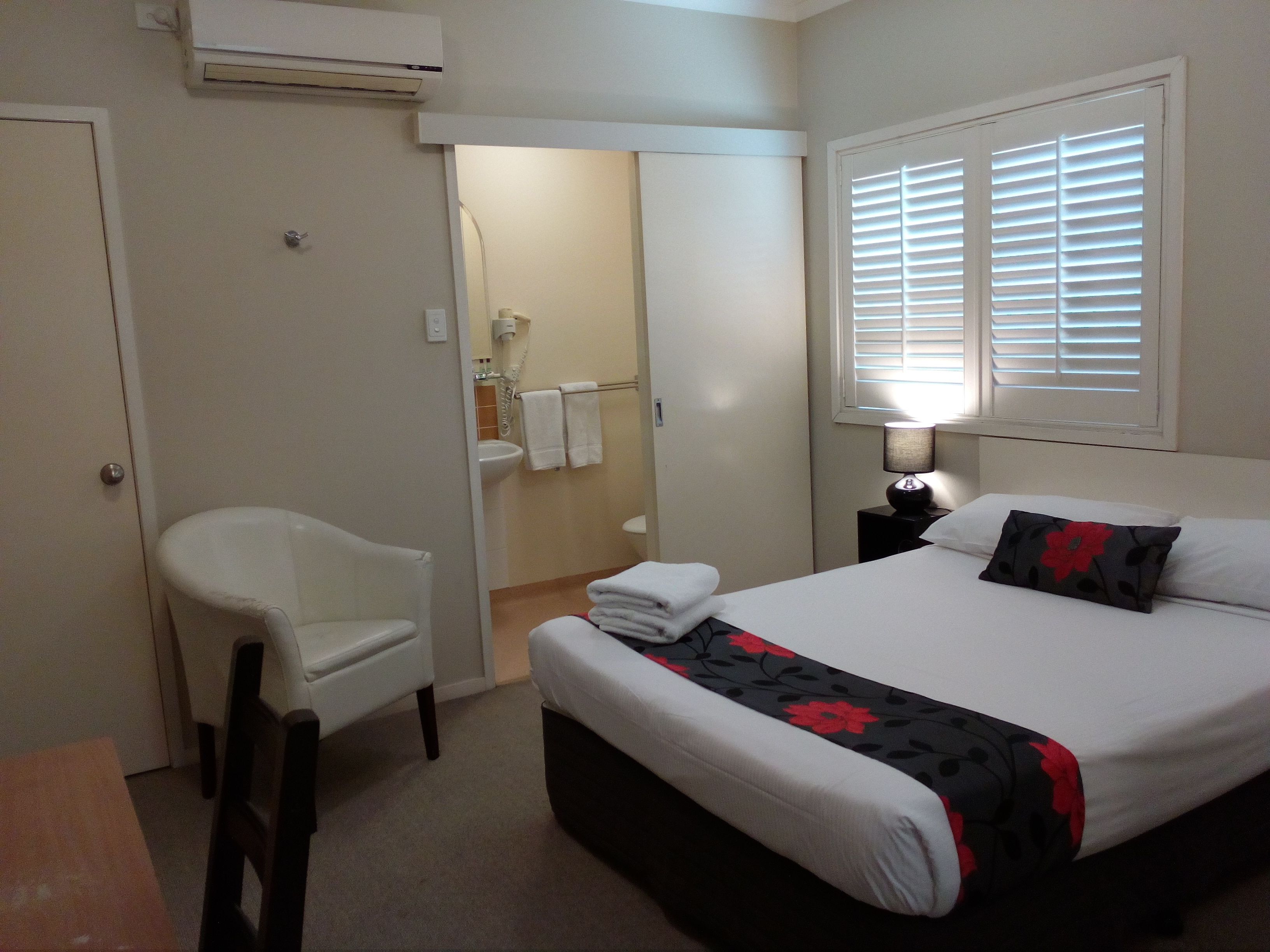 A hotel room with a bed, a chair, a bedside table with a lamp, a window with blinds, and an air conditioning unit.