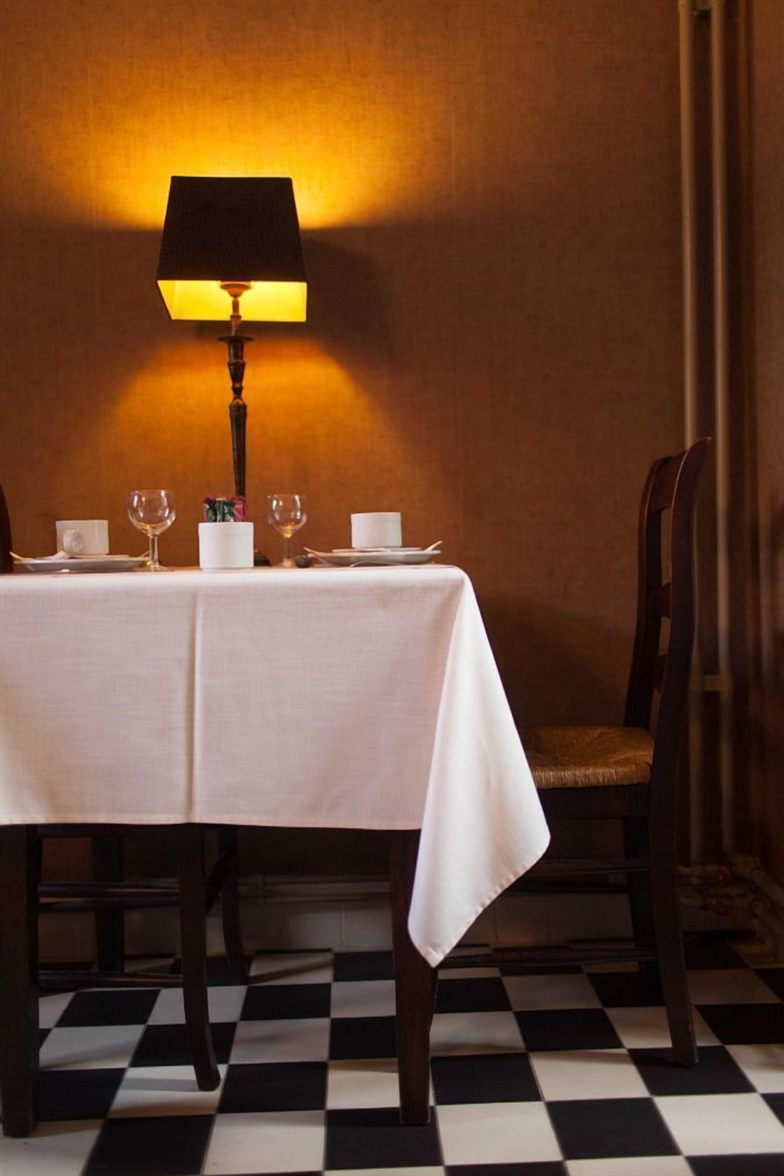 A dining table set with white tablecloth, glasses, and plates, with a lamp providing warm lighting.