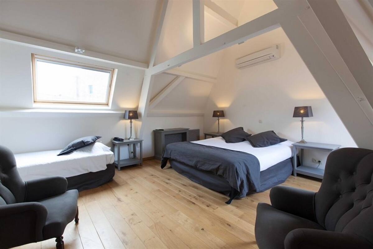 A small, cozy room with a loft bed, a single bed, two bedside tables with lamps, a chair, and a window.