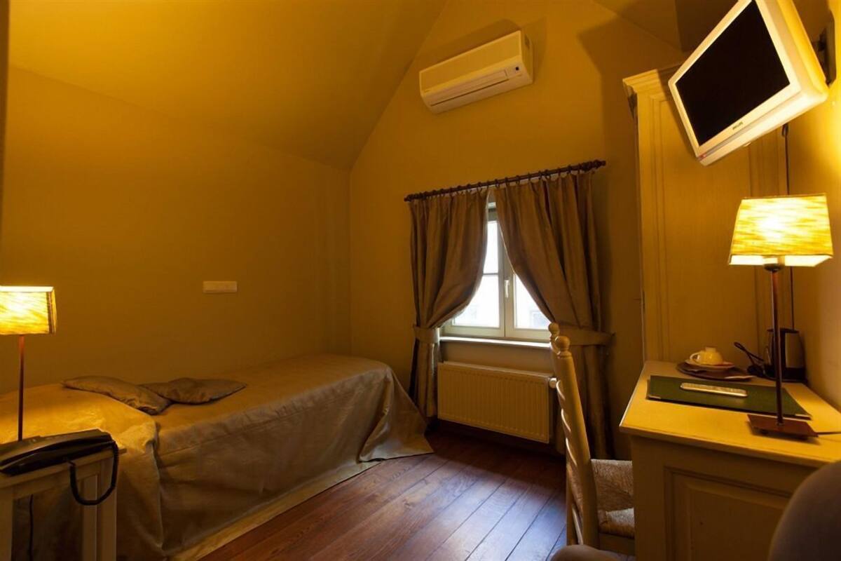 A small, cozy room with a single bed, a desk with a lamp, a television mounted on the wall, and a window with curtains.