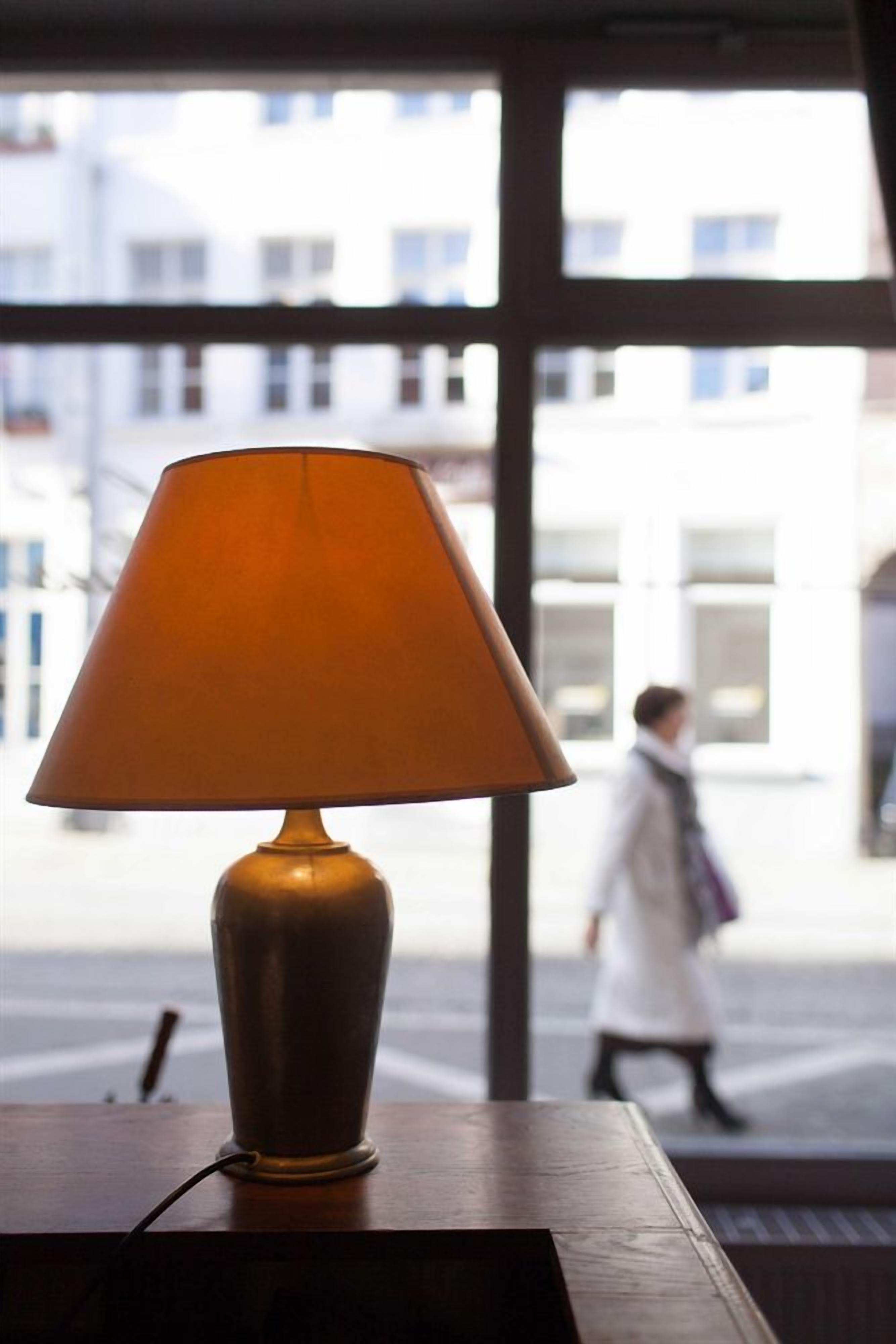 Une lampe posée sur une table près d’une fenêtre, d’où l’on peut voir des immeubles et des piétons.