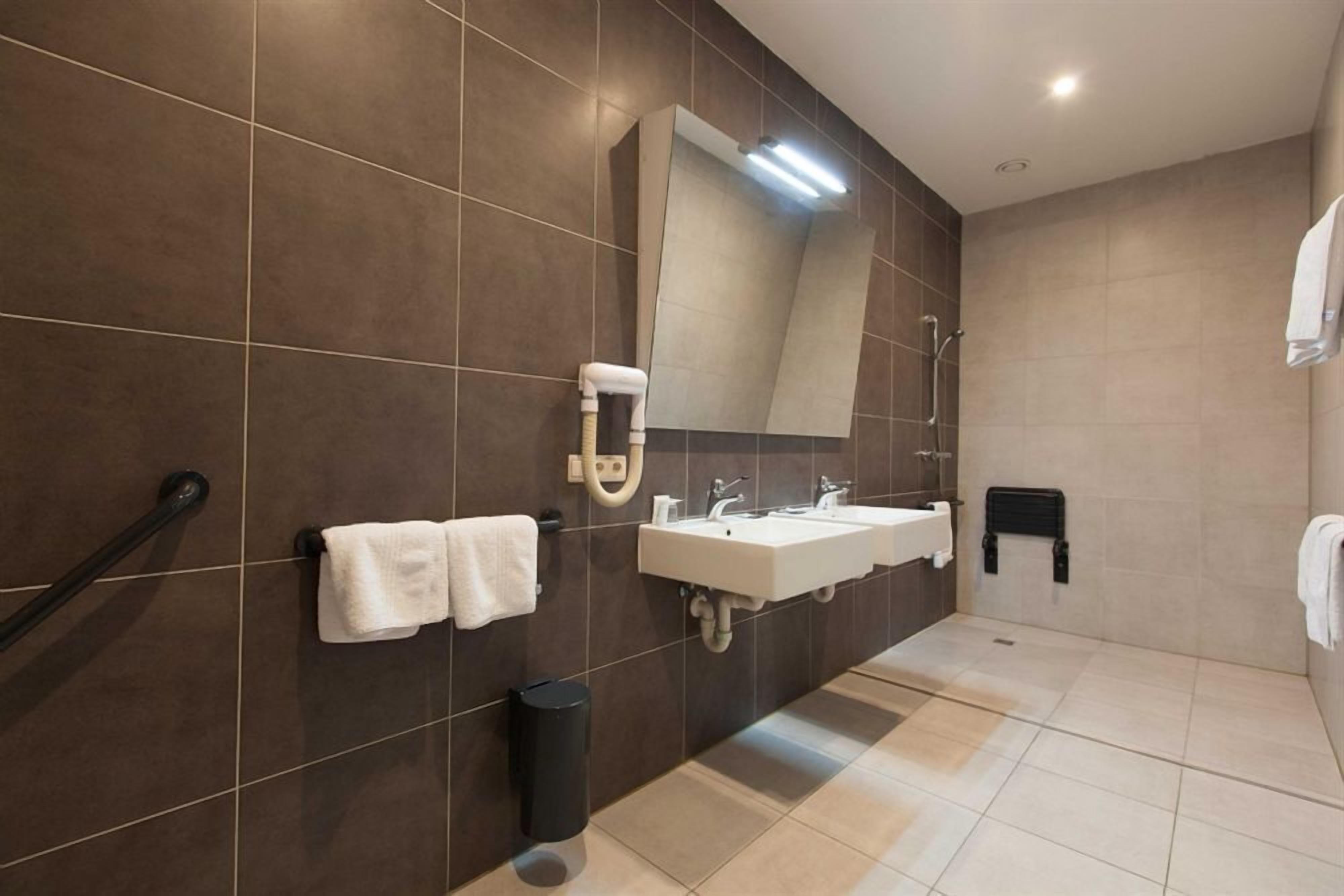 Um banheiro moderno com azulejos marrom-escuros, um espelho grande, duas pias, um porta-toalhas, uma lixeira e uma área de chuveiro.