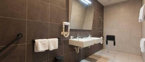 A modern bathroom with dark brown tiles, a large mirror, two sinks, a towel rack, a trash bin, and a shower area.