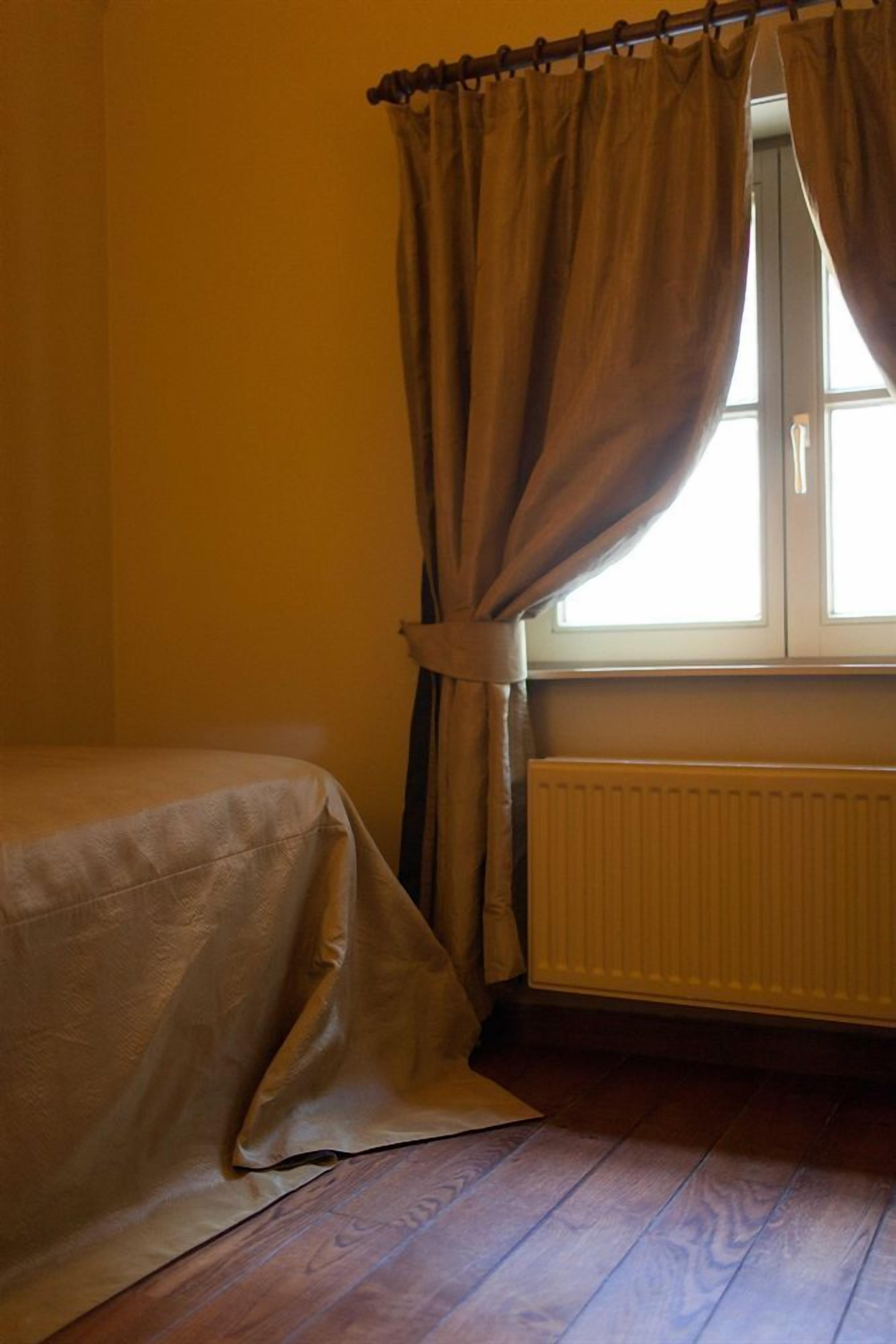 A room with a bed, a window with curtains, a radiator, and wooden flooring.