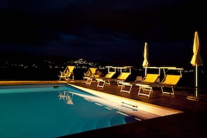 Un área de piscina nocturna con sillones de playa y sombrillas.