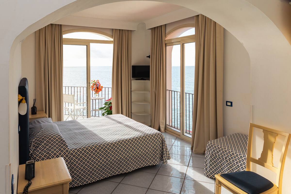 Habitación de hotel con cama, televisor, sillón, una mesita con un florero y balcón con vista al mar.