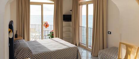 A hotel room with a bed, a TV, a chair, a small table with a vase of flowers, and a balcony with a view of the sea.