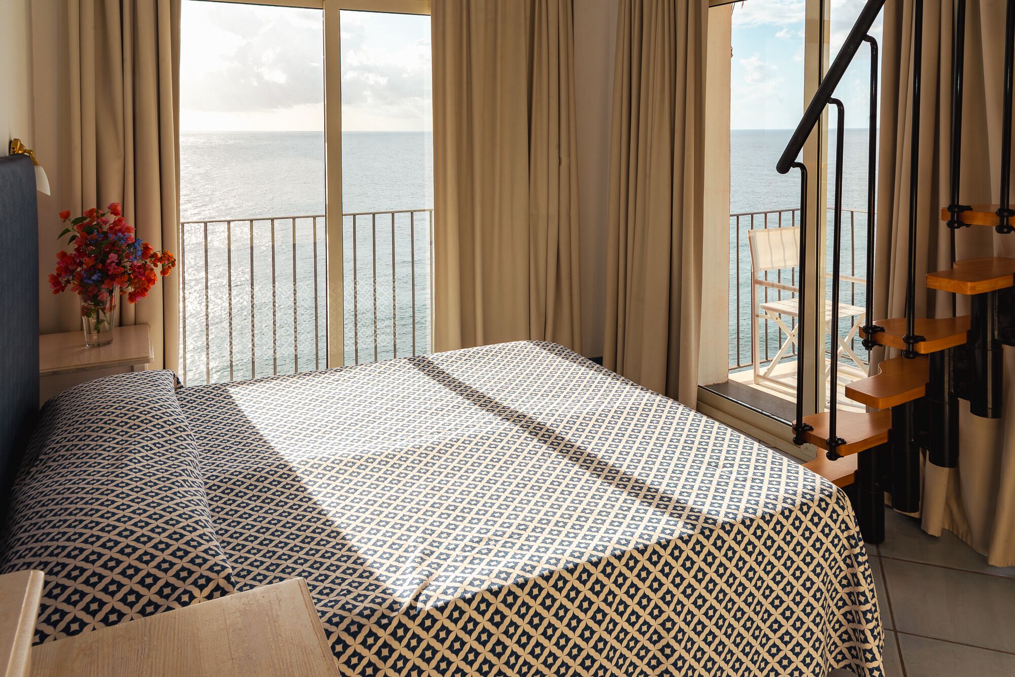 A bedroom with a bed, a window with curtains, a view of the sea, and a chair outside.