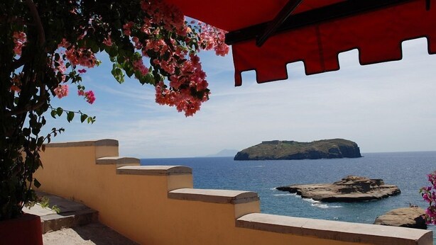 Vista costera con un muro de piedra y un toldo rojo, con vista al mar y una isla distante.