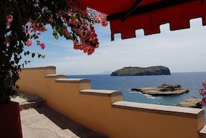 Vista costeira com muro de pedra e toldo vermelho, com vista para o mar e uma ilha ao longe.