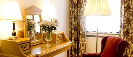 A room with a wooden desk, two lamps, a mirror, a vase with flowers, a red chair, and patterned curtains.