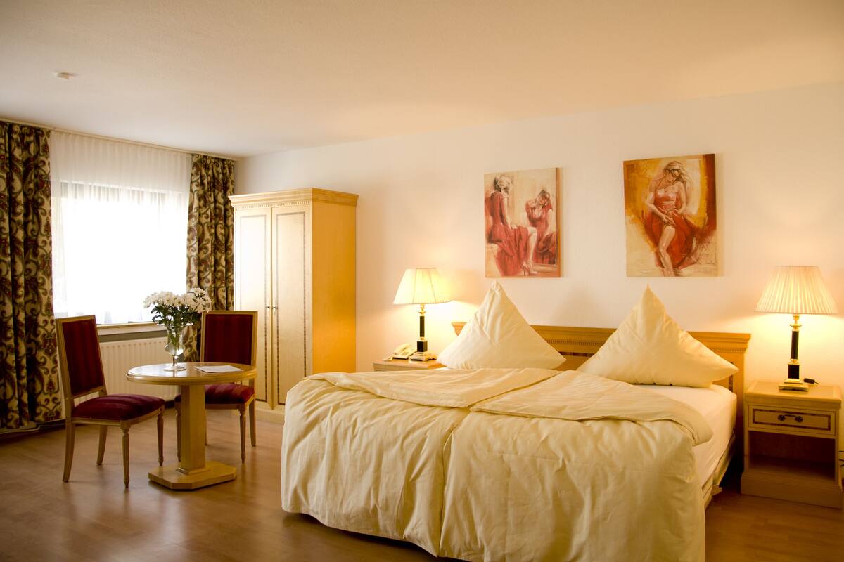 A hotel room with a bed, two bedside tables with lamps, a nightstand, a chair, a table with a vase of flowers, and a wardrobe.