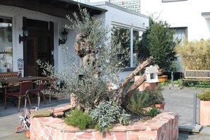 A stone planter with various plants, a wooden bench, and a table with chairs outside a building. - Hotel Kölner Hof Refrath (Bergisch Gladbach)