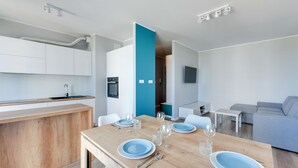 Ett modernt kök med vita skåp, ett träbord med blå tallrikar och glas, en grå soffa och en tv som är monterad på väggen.