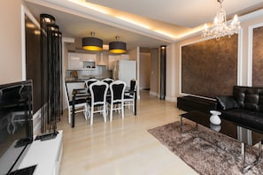 A modern living room with a black sofa, a coffee table, and a TV. There is a dining area with a table and chairs, and a kitchen in the background.