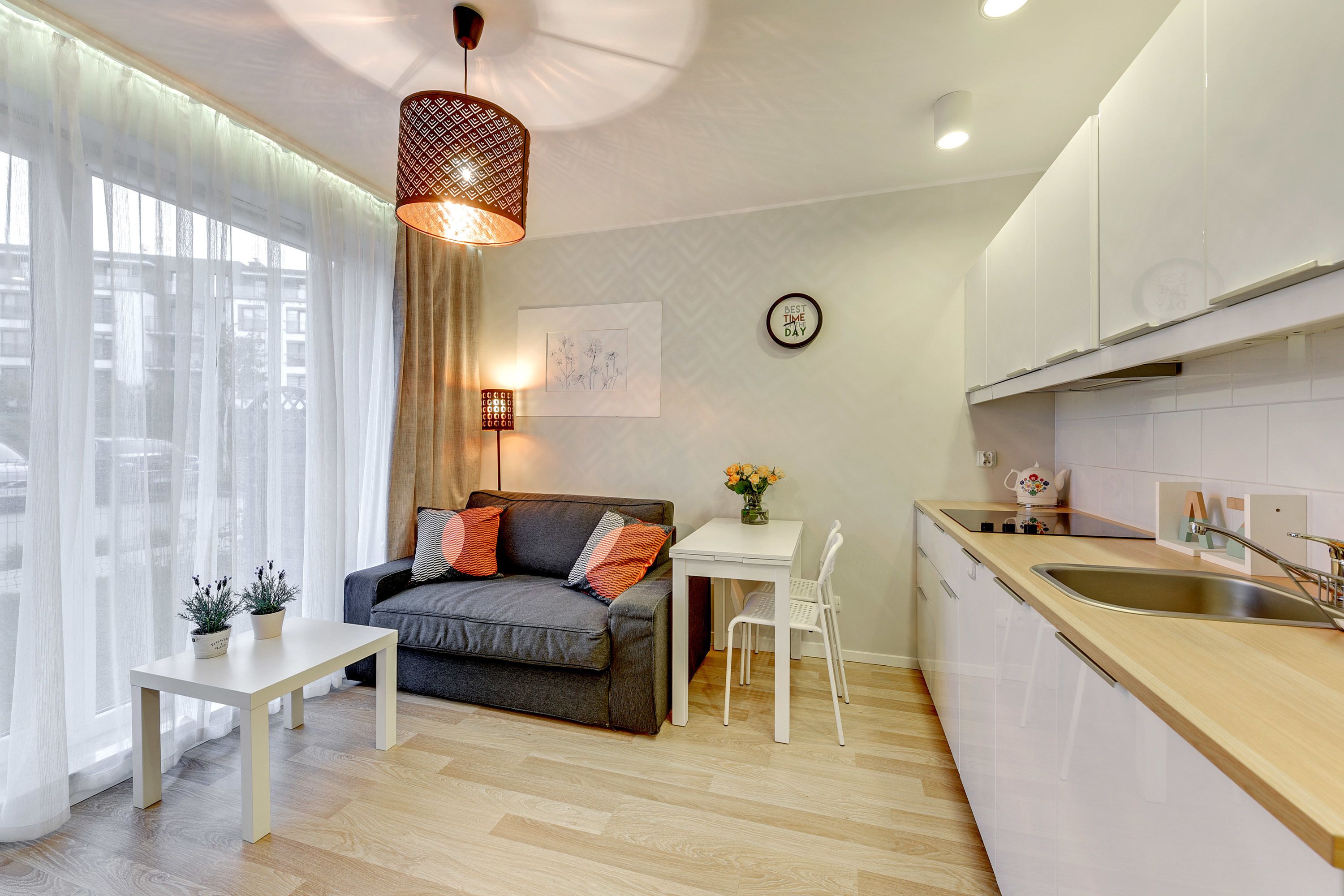 A modern kitchen with white cabinets, a wooden countertop, and a sink. There's a dining area with a white table and chairs, and a sofa with cushions.