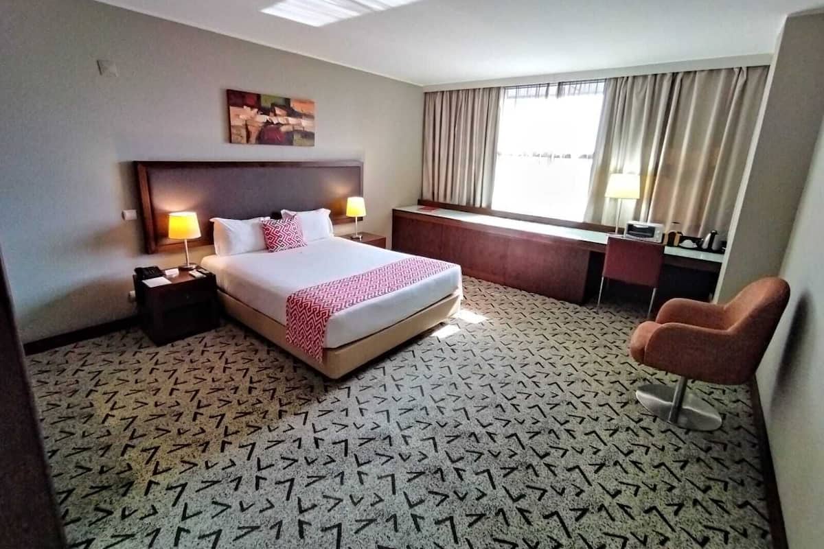 A hotel room with a bed, bedside tables, a desk, a chair, a window with curtains, and a patterned carpet.