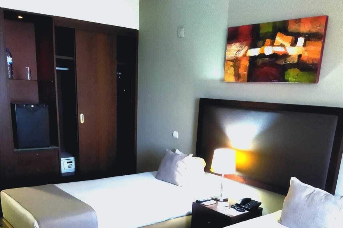 A hotel room with two single beds, a nightstand with a lamp, a telephone, and a painting on the wall.
