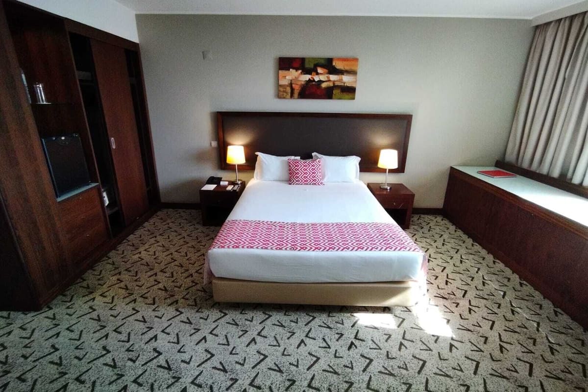 A hotel room with a large bed, two bedside tables with lamps, a desk with a chair, a wardrobe, and a painting on the wall.