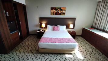 A hotel room with a large bed, two bedside tables with lamps, a desk with a chair, a wardrobe, and a painting on the wall.
