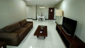 A living room with a brown sofa, a coffee table with a vase, a TV on a stand, and a dining area in the background. - Skyna Hotel Luanda (Luanda)