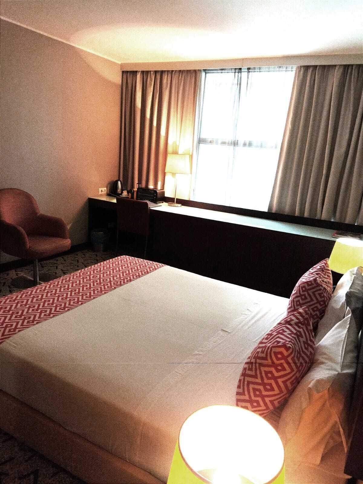 A hotel room with a large bed, a desk, a chair, and a window with curtains.