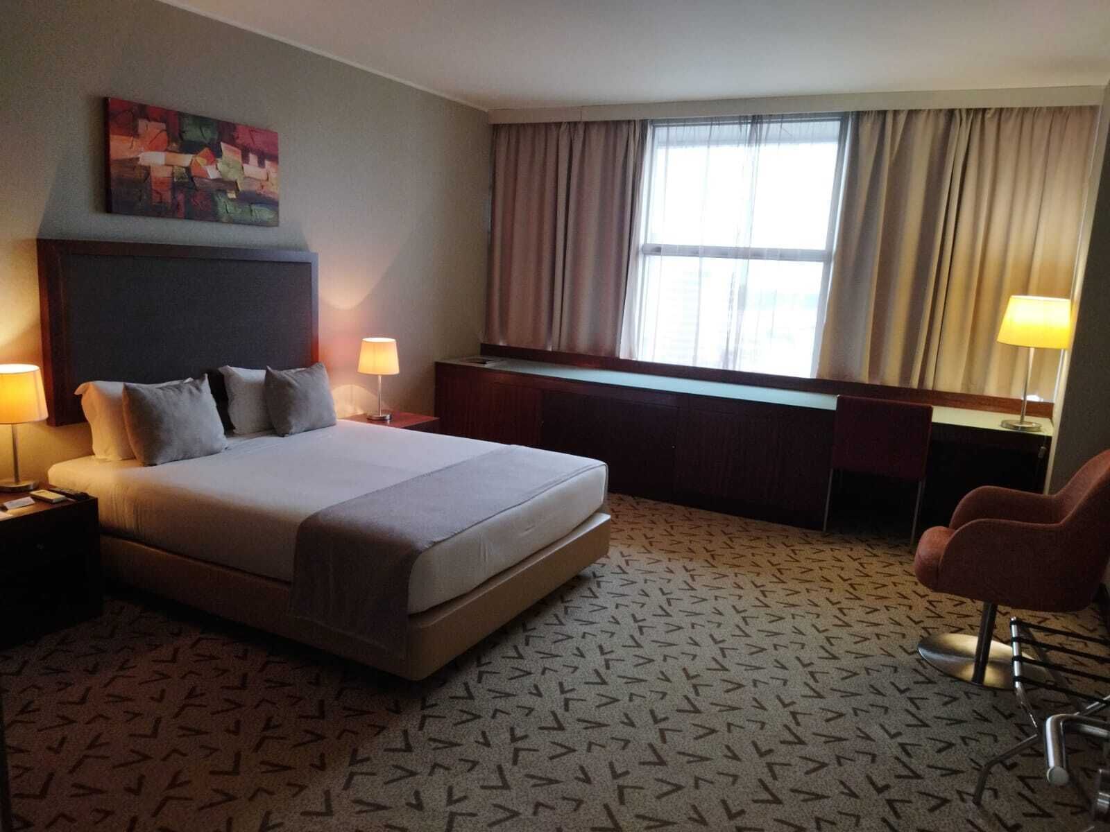 A hotel room with a large bed, a desk, a chair, a window with curtains, and a painting on the wall.