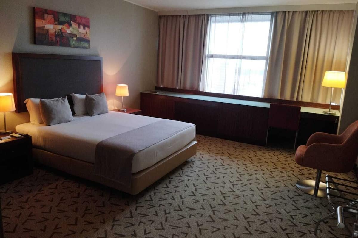 A hotel room with a large bed, a desk, a chair, a window with curtains, and a painting on the wall.