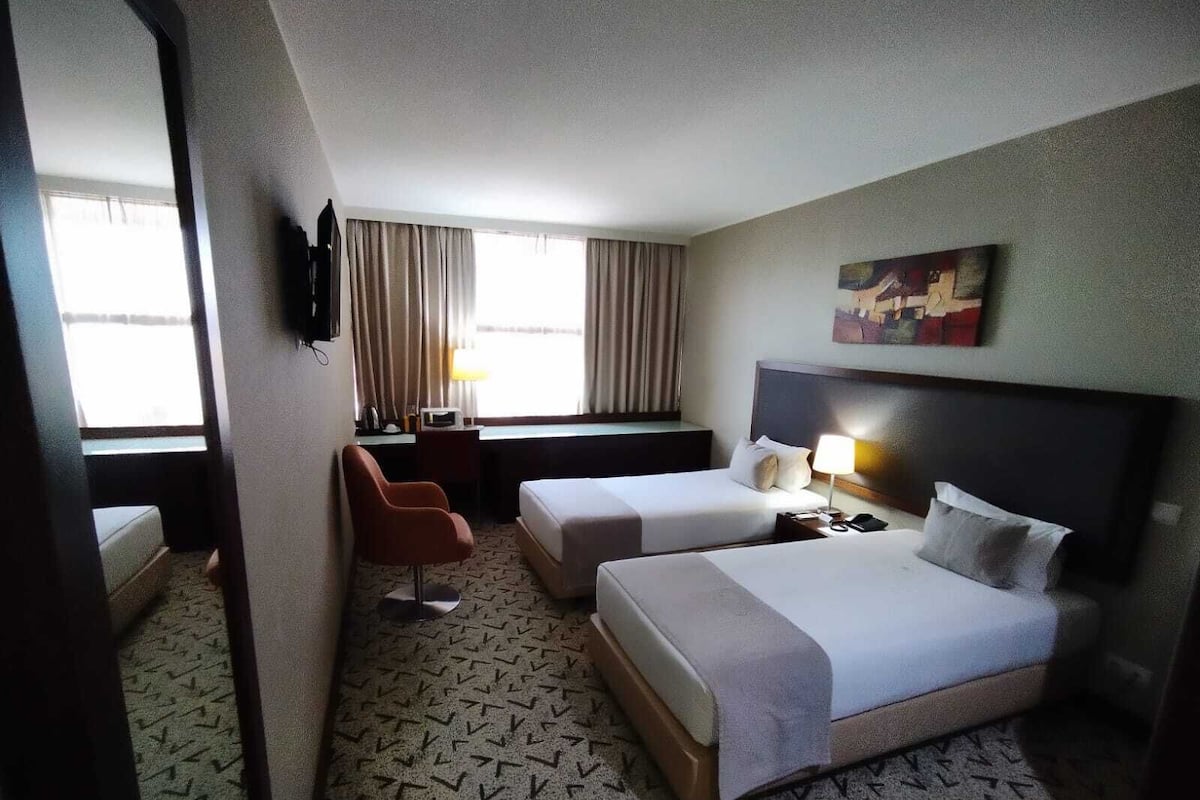A hotel room with two beds, a desk, a chair, a TV, and a painting on the wall.