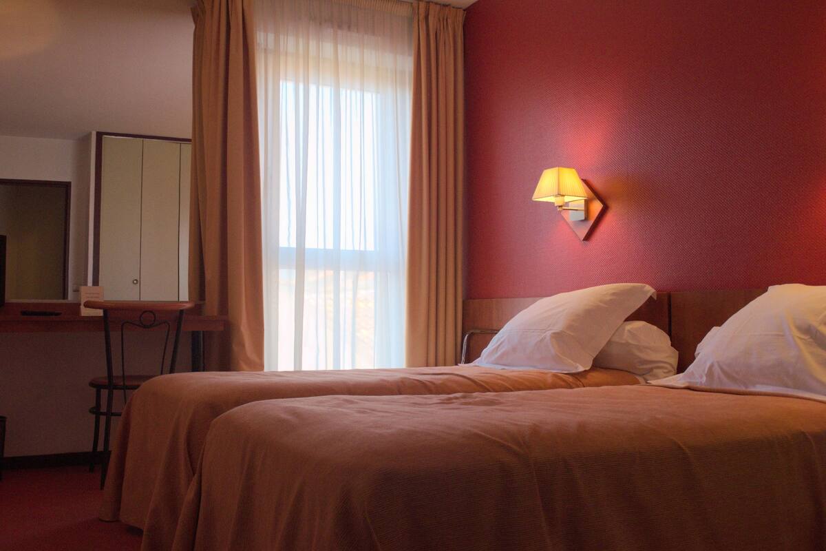 A hotel room with two beds, a desk with a chair, a mirror, and a window with curtains.